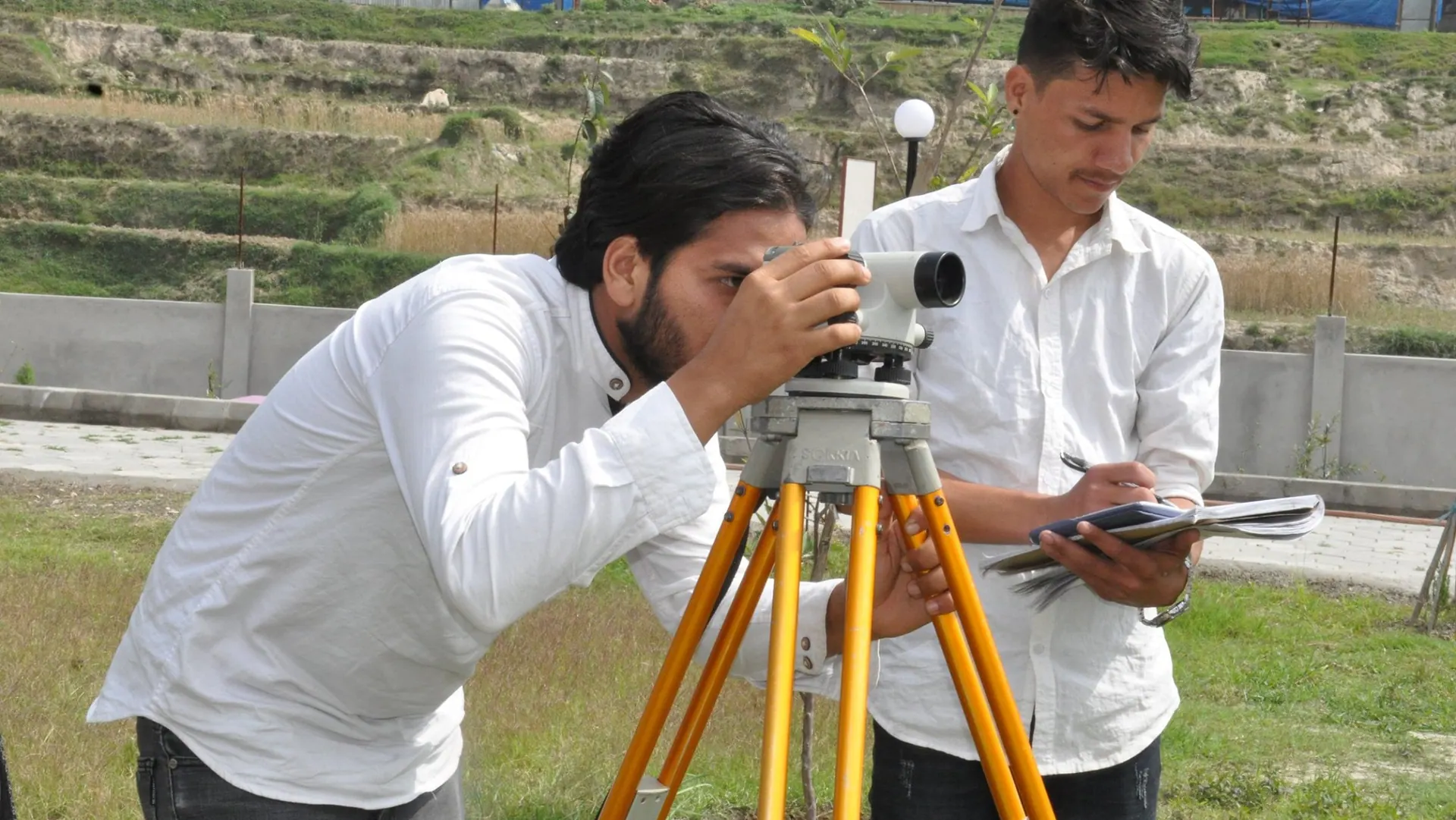 A civil engineering student sighting through a levelling instrument while a classmate records the reading